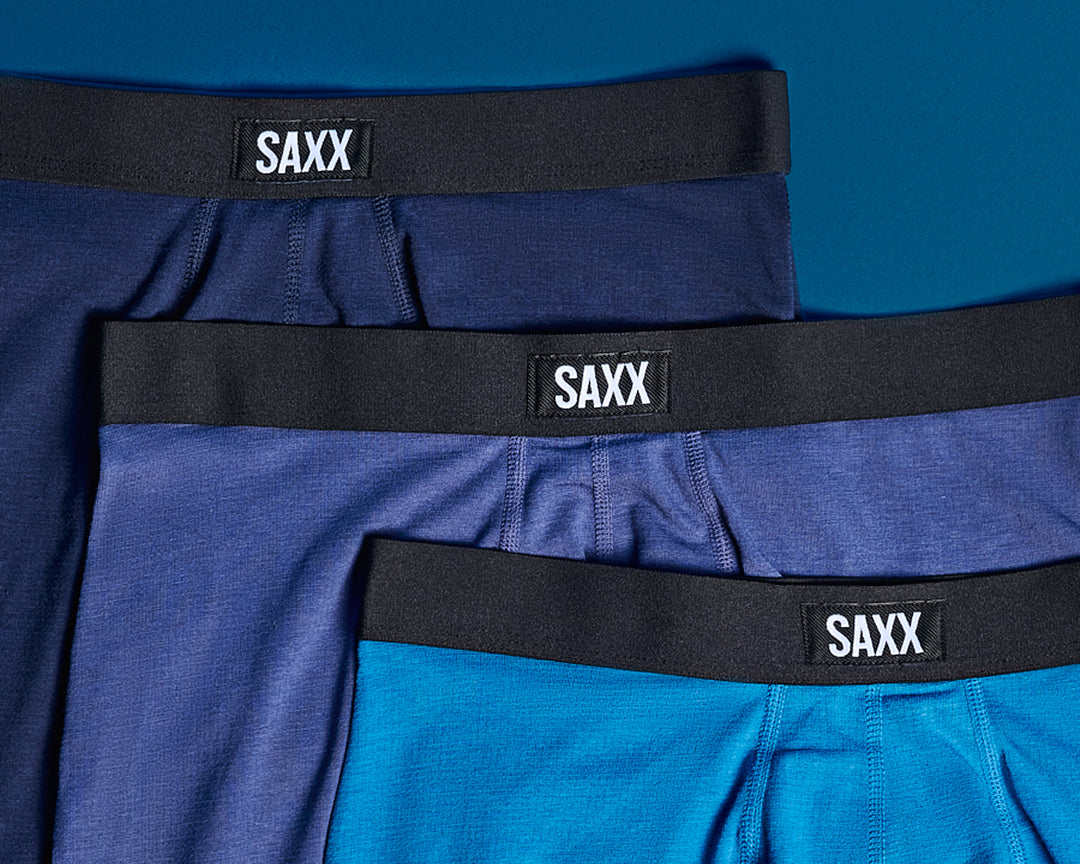 Three pairs of blue SAXX underwear with black waistbands on a blue background.