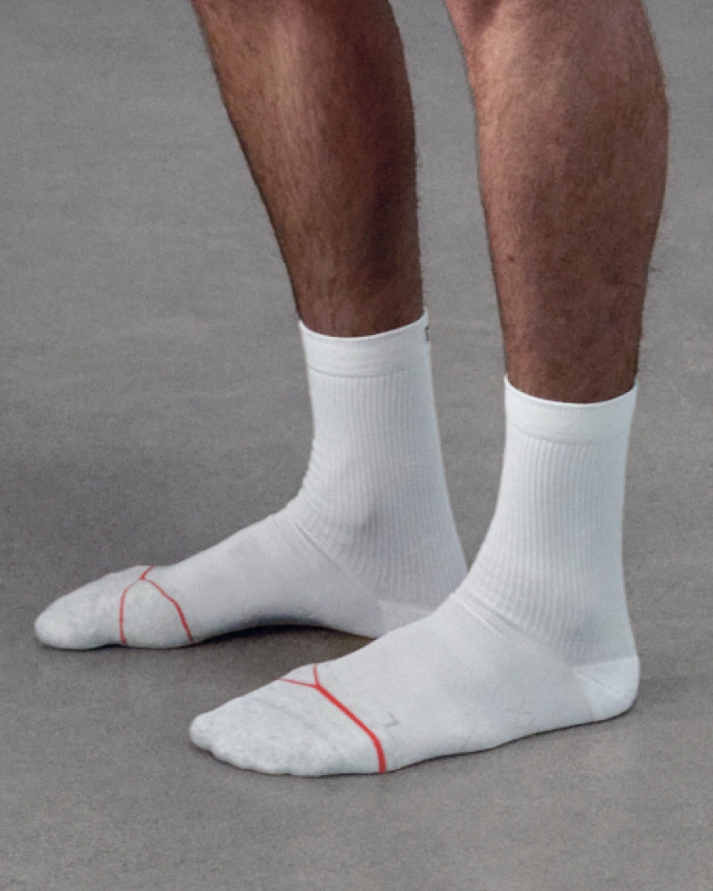 White crew socks with thin red accent lines on the toes, worn on a person's feet against a gray floor.