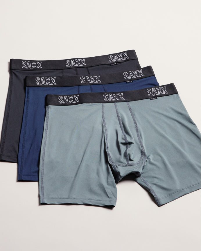 Three pairs of SAXX men's boxer briefs in dark gray, navy blue, and light gray, arranged in a fan shape on a light beige background.