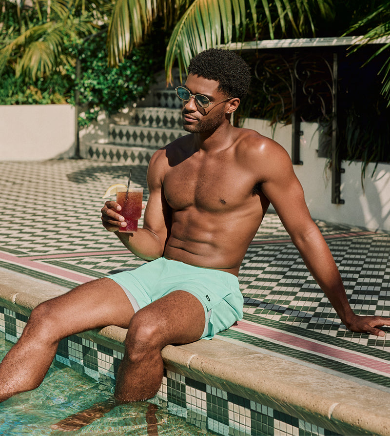 Man in swim trunks sitting with his feet dangling in pool while holding a drink